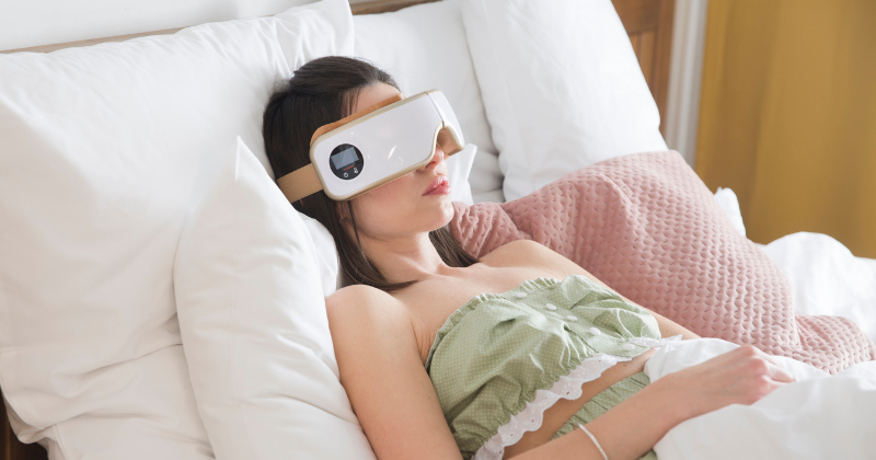 A woman using an eye massager while lying in bed.